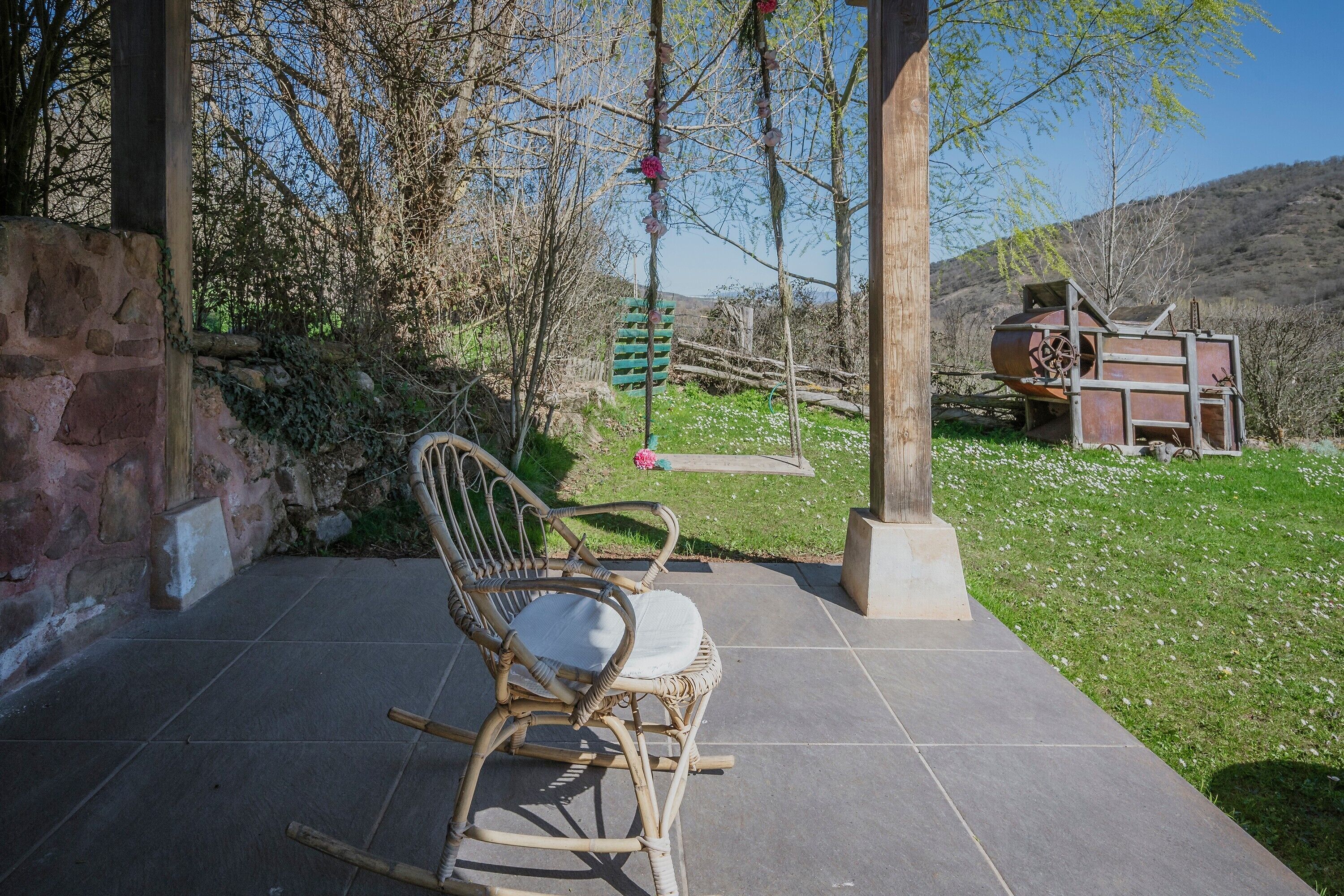 Un patio con sillas de mimbre, un muro de piedra y un espacio exterior rústico con una estructura verde y un objeto metálico grande.
