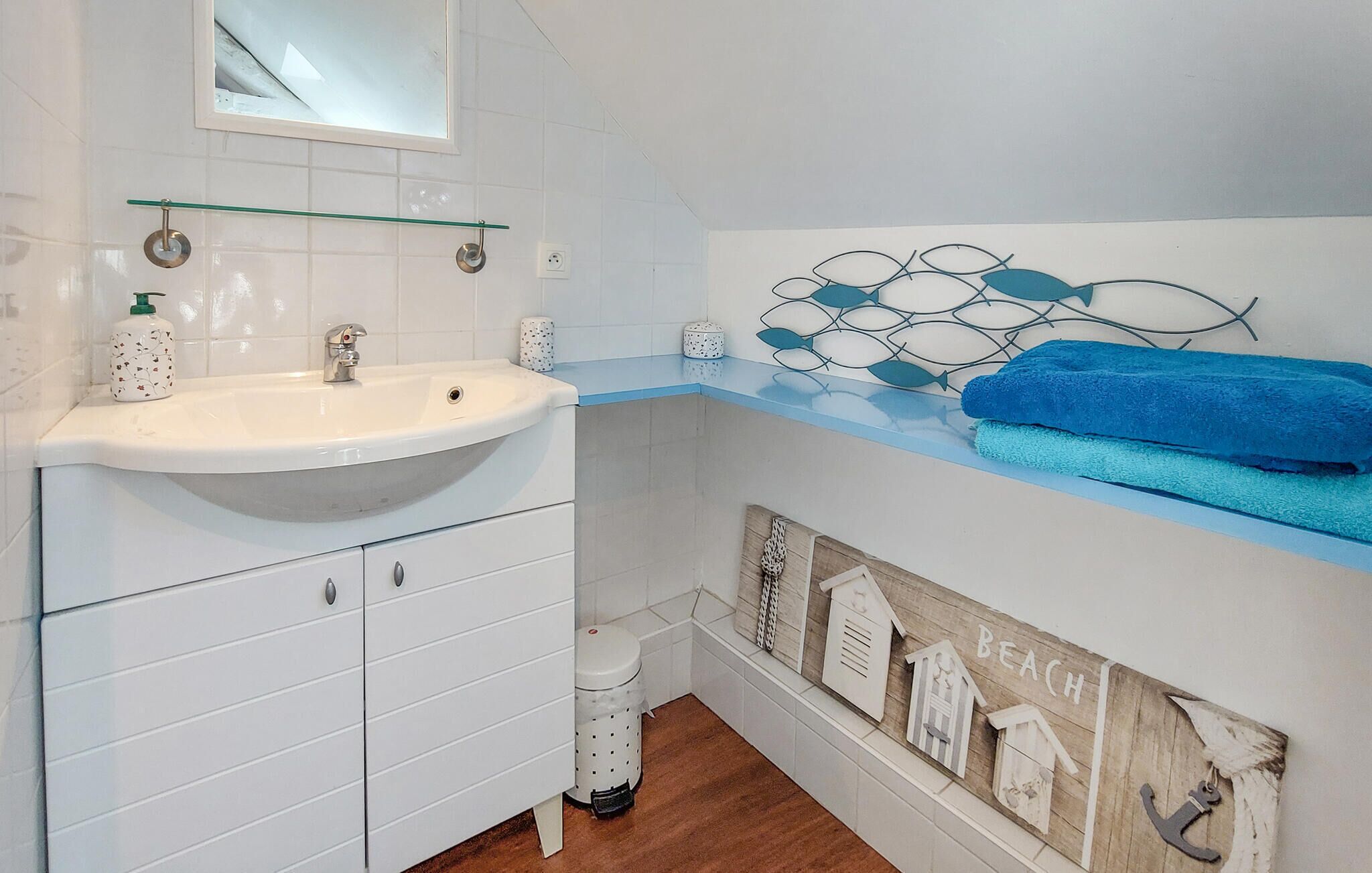 A bathroom with a white sink, a mirror above it, and a towel rack with blue towels. There is a decorative wall piece with a beach theme and a small trash bin.