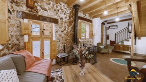 Un intérieur rustique avec des murs en pierres apparentes, des poutres en bois, un coin salon confortable avec un canapé et des coussins, une table en bois ornée d’un vase, et un escalier menant à un autre niveau.