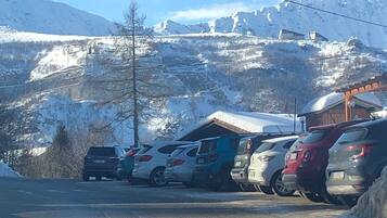 Ein Parkplatz mit mehreren Autos, ein Gebäude mit Satteldach und verschneite Berge im Hintergrund.