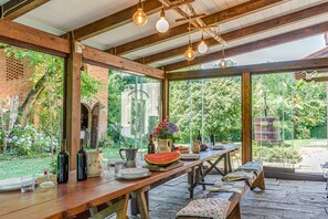 Un espacio exterior techado para comer con una mesa de madera, lámparas colgantes y vistas a un muro de ladrillo y vegetación.
