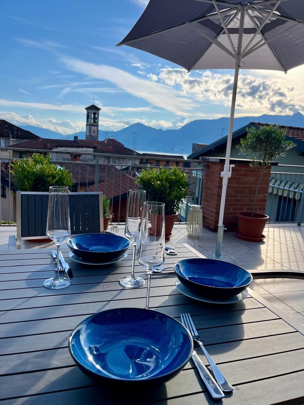 Une table dressée en plein air, avec de l’argenterie bleue, des verres à vin et des couverts, posée sur une table en bois, sous un grand parasol, avec une vue sur les montagnes en arrière-plan.