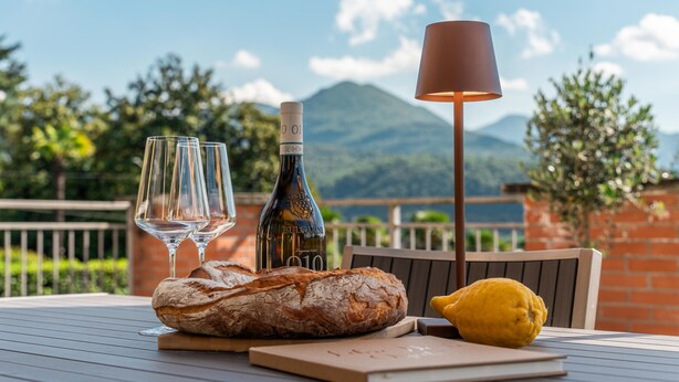 Une table avec une bouteille de vin, deux verres, un pain, un citron et une lampe, le tout disposé devant un paysage montagneux pittoresque.