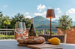 Ein Tisch mit einer Weinflasche, zwei Gläsern, einem Brotlaib, einer Zitrone und einer Lampe, vor einer malerischen Berglandschaft.