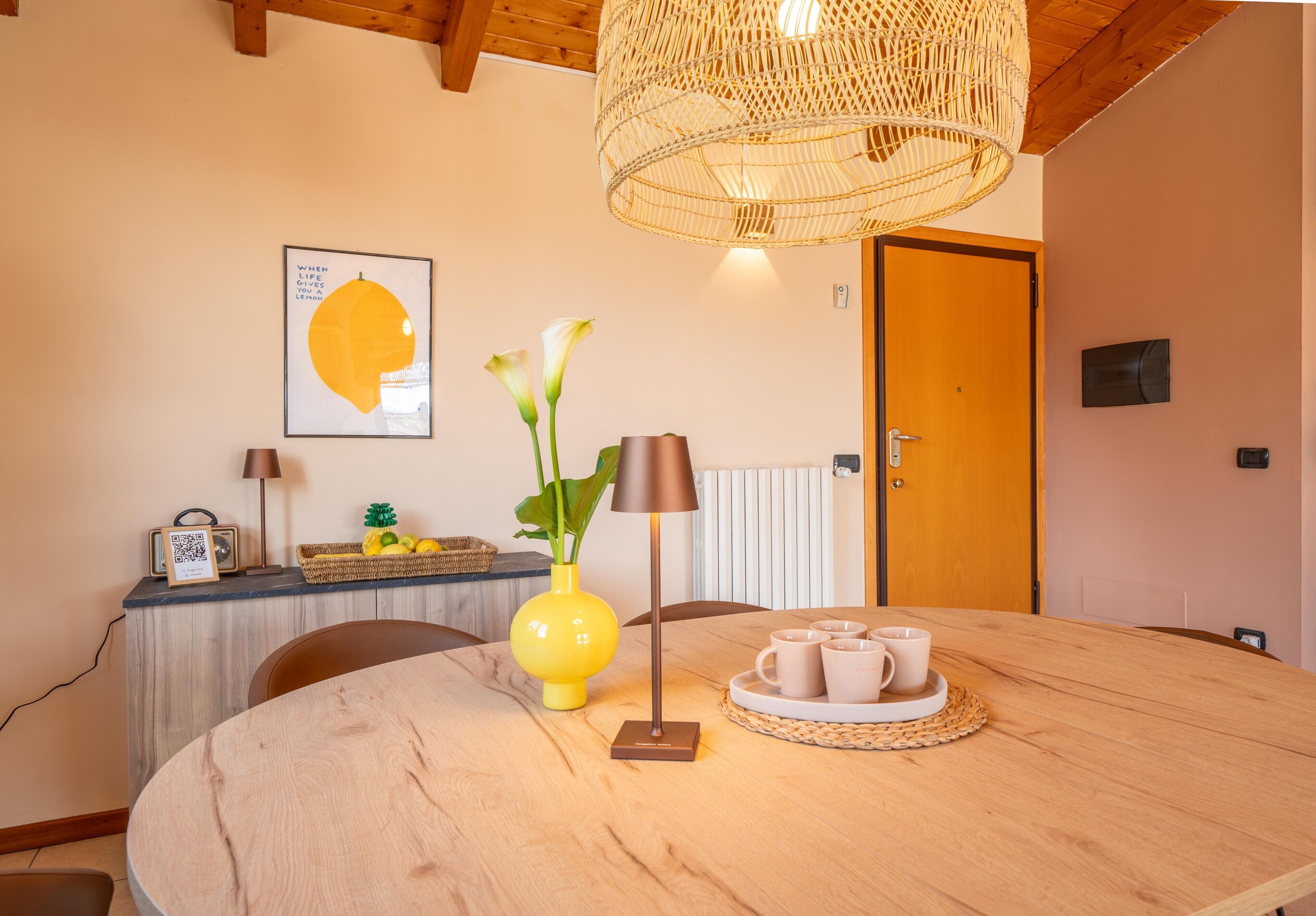 Un coin repas avec une table ronde en bois, un vase jaune orné de fleurs, une lampe et un plateau avec des tasses.