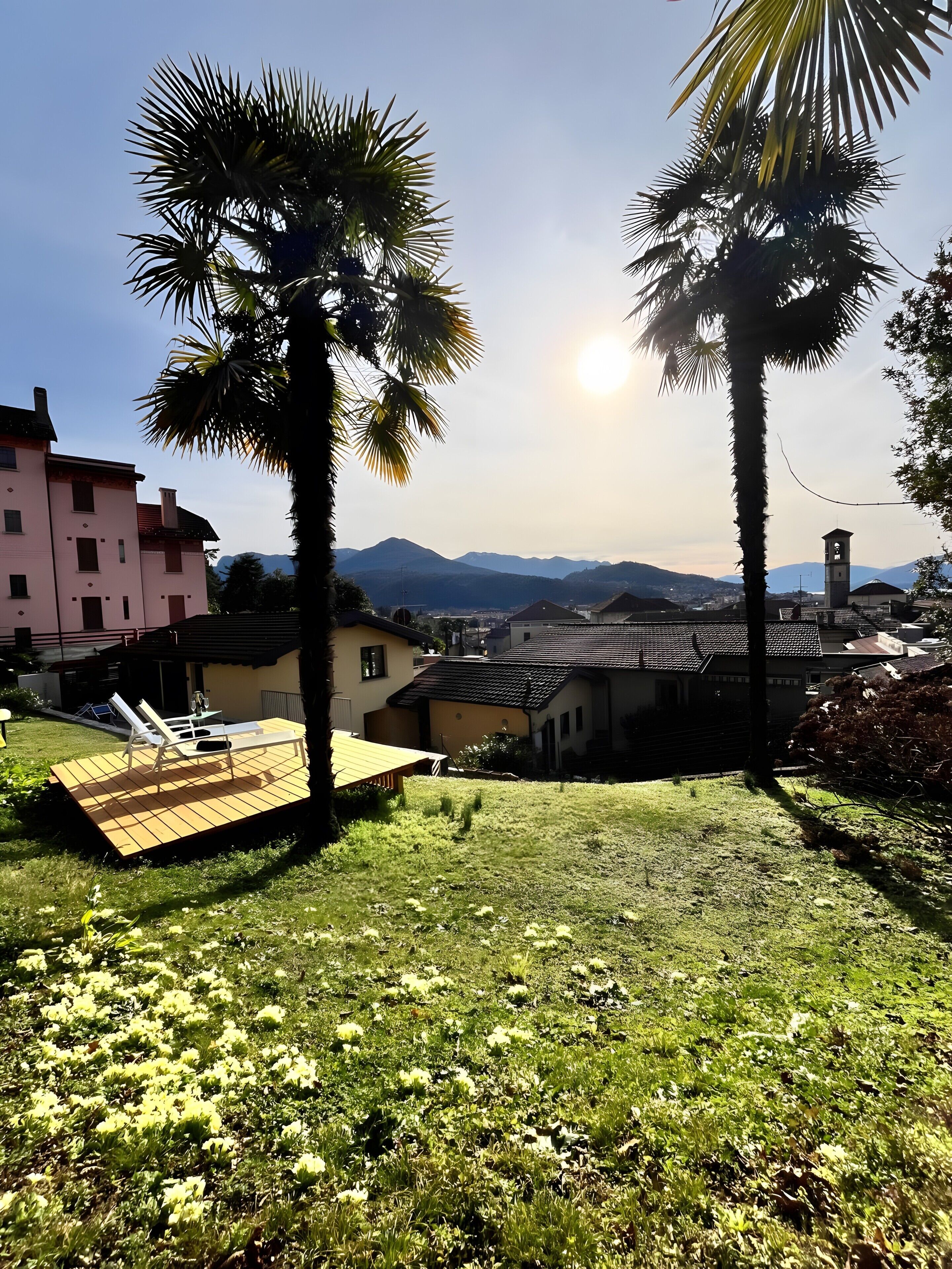 Un espace verdoyant avec des palmiers, une terrasse en bois dotée de chaises longues et une vue sur des bâtiments et des montagnes en arrière-plan.