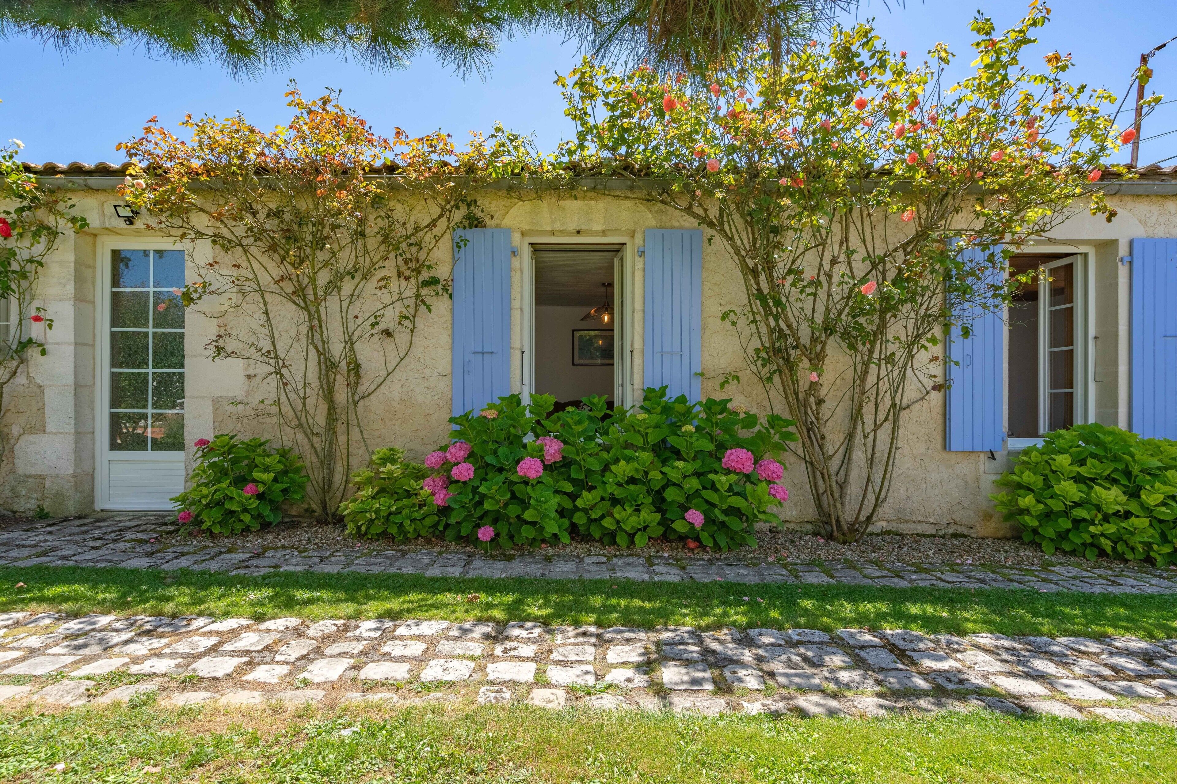 Un bâtiment à la façade en pierre, avec des volets bleus et un jardin où poussent des fleurs roses.