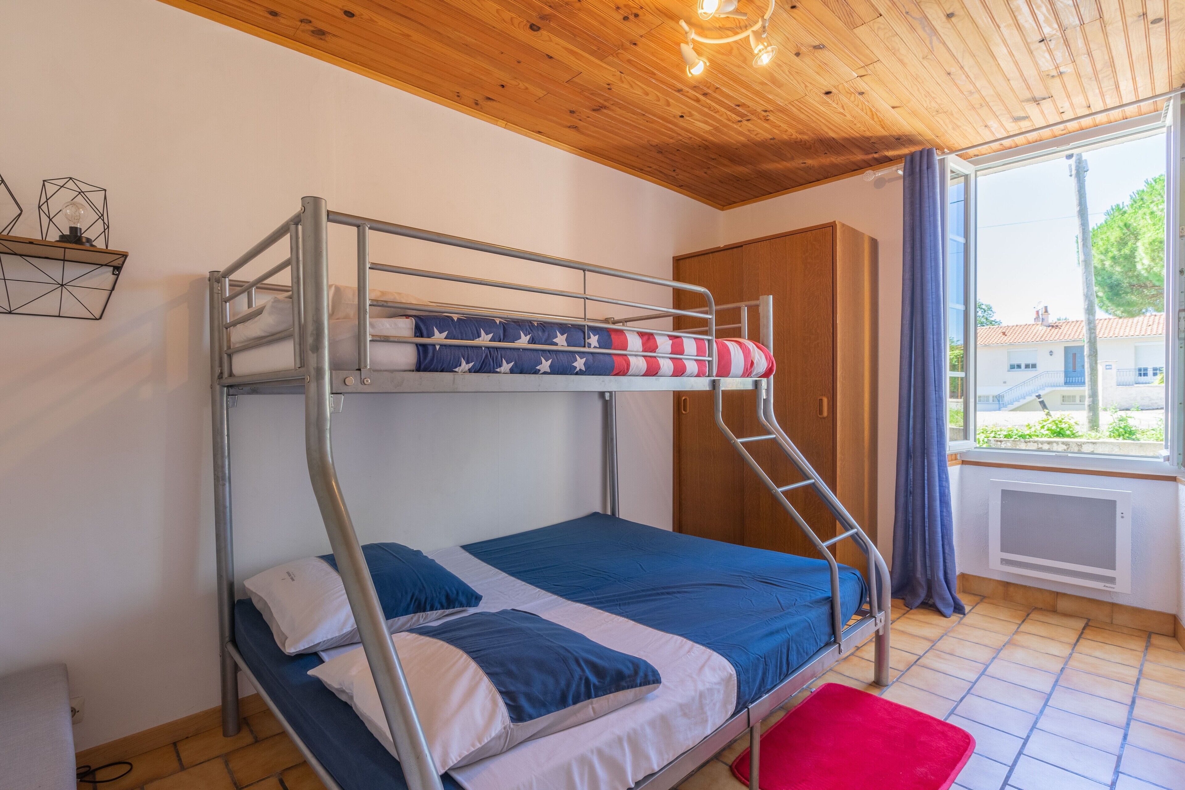 A bunk bed room with a wooden ceiling, a window with curtains, and a view of the outside.