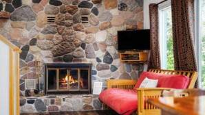 Une cheminée en pierre avec un feu de bois, un téléviseur fixé au mur, un canapé rouge et une table en bois.