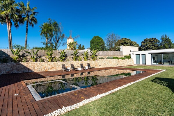 A swimming pool with a wooden deck, surrounded by a stone wall and lush greenery.