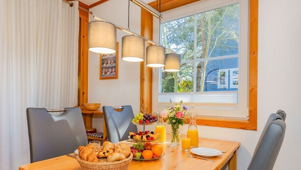 Una mesa de comedor con comida para el desayuno, sillas, una ventana con vistas y una lámpara colgante.