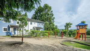Un parque infantil con un tobogán y estructuras para escalar, rodeado de arena y follaje, con casas al fondo.