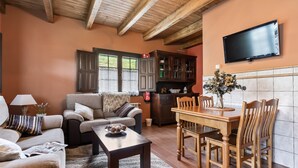 Ein Wohnzimmer mit einer Couch, einem Couchtisch und einem Esstisch mit Stühlen. Ein Fernseher ist an der Wand montiert und es gibt eine Küchenzeile mit Schränken und einer gefliesten Spritzwand.