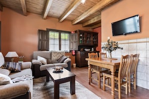Una sala de estar con sofá, mesa de centro y mesa de comedor con sillas. Hay un televisor montado en la pared y una cocina con armarios y salpicadero de azulejos.