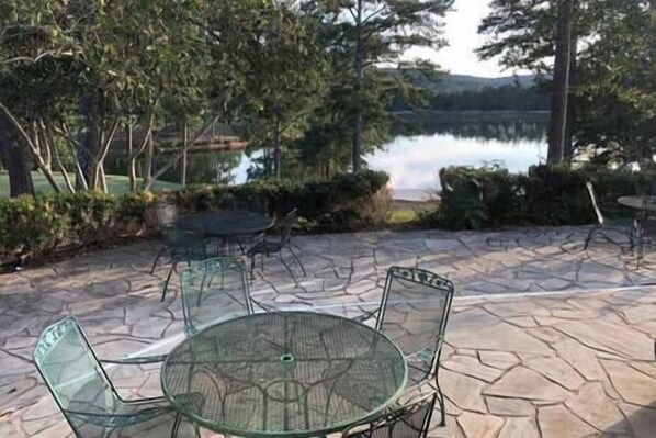 Un patio al aire libre con una mesa redonda de vidrio y sillas, piso de piedra y vista a un lago.