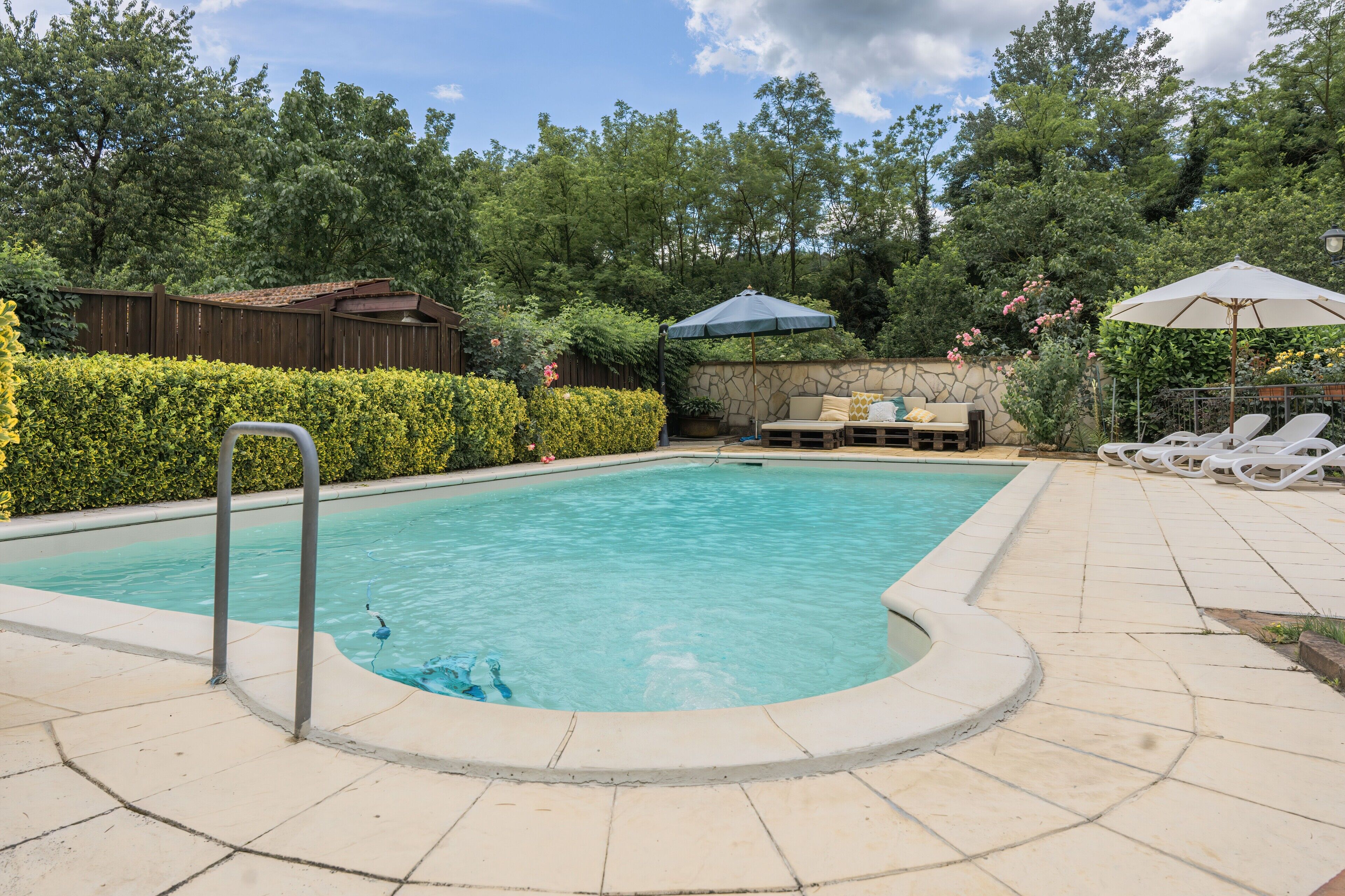 Une piscine bordée d’un muret en pierre, une clôture en bois et un espace de détente agrémenté de parasols.