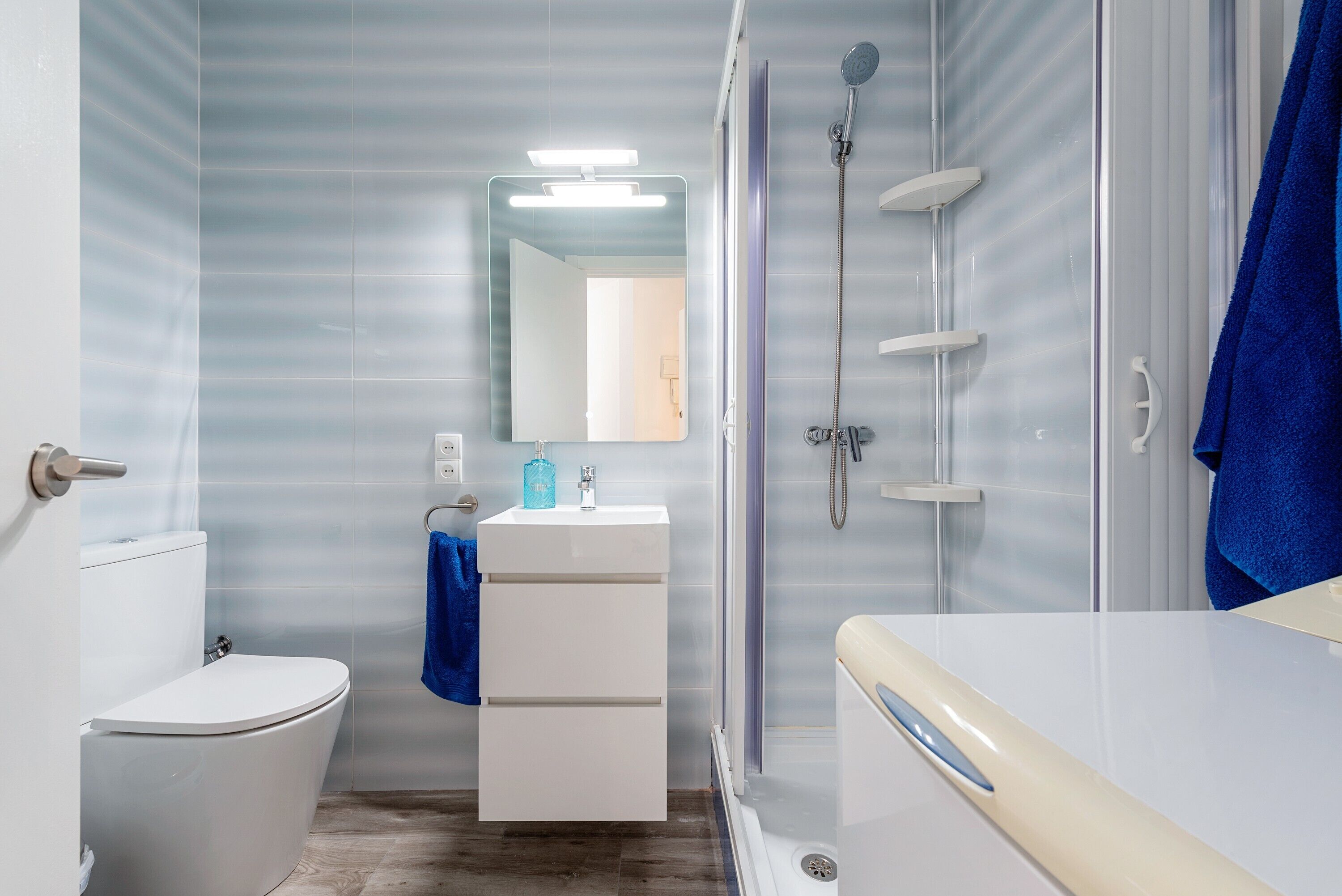 Un baño con inodoro, lavamanos con espejo, ducha y una toalla azul.