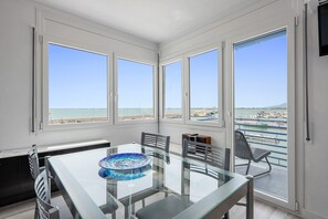Quarto moderno com mesa de vidro, cadeiras e grandes janelas com vista para a praia e o porto de iates.