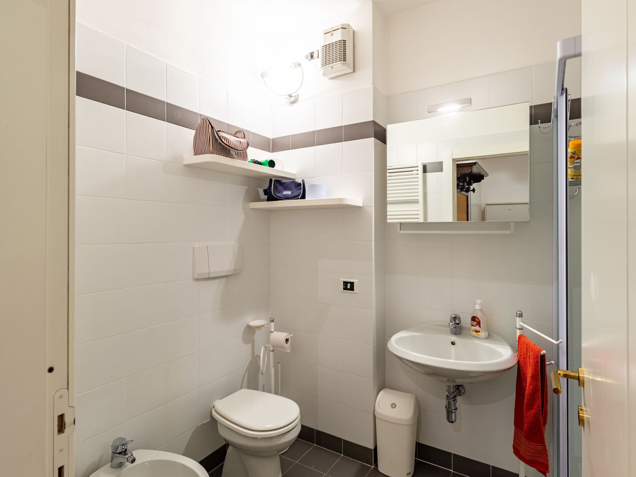 A bathroom with a toilet, sink, and a mirror. There are shelves above the toilet and a towel hanging on the door.