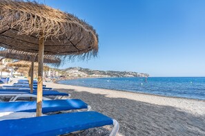 A beach with blue lounge chairs and a straw umbrella. - Apartment 'Primera Linea De Playa La Herradura' with Sea Views, Balcony and Wi-Fi (Almuñécar)