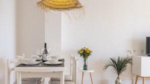 Een eetgedeelte met een witte tafel, stoelen, een geweven lamp, een vaas met gele bloemen en een potplant.