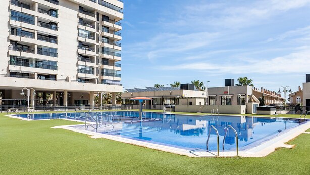A swimming pool with a surrounding grassy area, a multi-story building in the background, and a clear sky. - Apartment 'Valencia Playa' with Sea View, Wi-Fi and Air Conditioning (Alboraya)