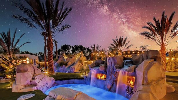 A nighttime outdoor area with a rock formation, water feature, and palm trees.