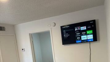 Una sala de estar moderna con un televisor de pantalla plana montado en la pared, piso de madera, una mesa de centro de vidrio y un jarrón, y una puerta que da a otra habitación.