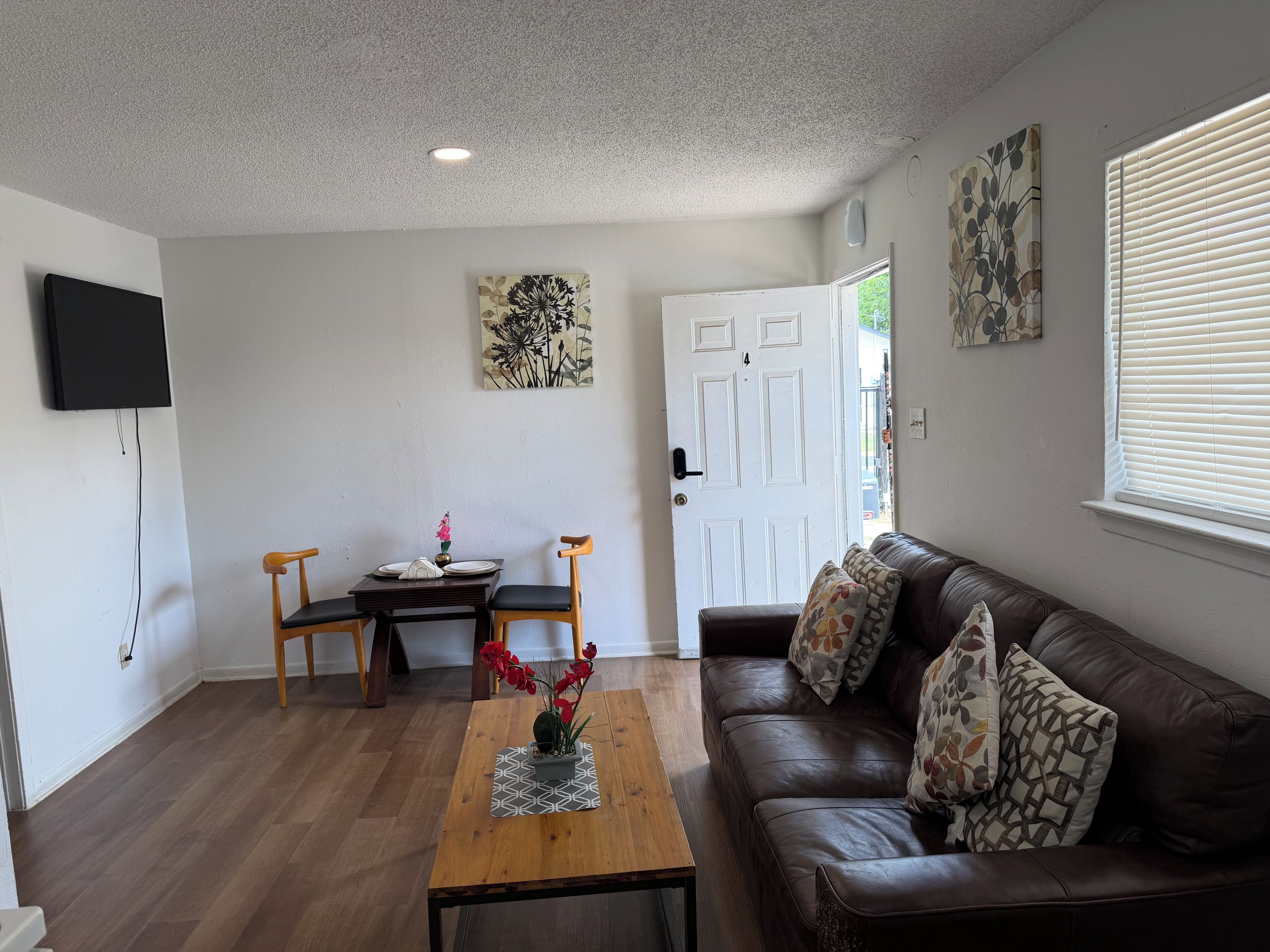 A living room with a brown leather sofa, a wooden coffee table, and a dining table with chairs. There is a TV mounted on the wall, and a door leading outside.