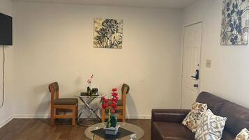 Una sala de estar con un sofá de cuero marrón, una mesa de centro de vidrio, un tapete rojo, dos sillas de madera, una mesita con flores y un televisor montado en la pared.