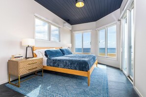 A bedroom with a bed, bedside tables, and a view of the sea through large windows.