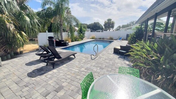 Une piscine entourée de chaises longues et agrémentée d’une table en verre, le tout bordé de plantes en pot et d’une clôture blanche.