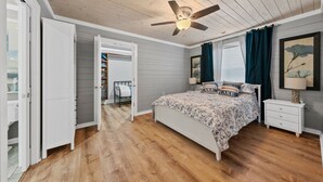 Une chambre avec un plafond en bois, un lit recouvert d’une literie à motifs floraux, une table de chevet, un ventilateur de plafond et une fenêtre avec des rideaux.
