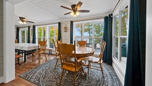 Un coin repas avec une table ronde et six chaises en bois, de grandes fenêtres avec des rideaux et un ventilateur de plafond.