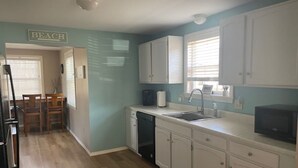 A compact kitchen with white cabinets, a sink, and a microwave. There is a dining area with a table and chairs visible through an open doorway. - Wells Charmer 2 Bed close to Wells Beach, Restaurants, AC, WiFi. 
Sleeps 6, Deck (Wells)