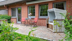 Une maison en briques avec un patio comprenant une table et des chaises, un fauteuil en osier blanc et un petit jardin.