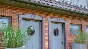 Un bâtiment à deux étages avec une façade en briques, deux portes grises dotées de fenêtres rondes et un toit recouvert de tuiles.