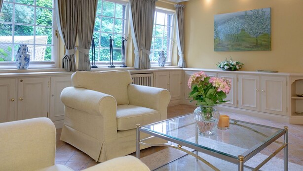 Un salon avec un fauteuil beige, une table basse en verre et un vase rempli de fleurs. On y trouve des fenêtres avec des rideaux, un tableau accroché au mur et un radiateur.
