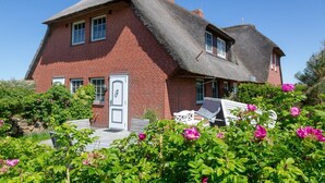 Ein rotes Backsteinhaus mit einem Reetdach, einer weißen Tür und einem Garten mit rosa Blumen.
