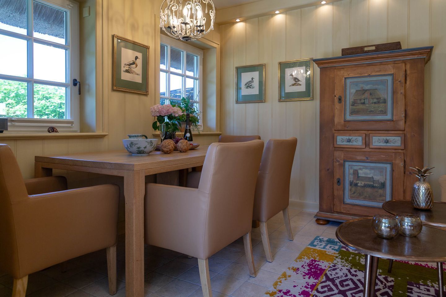 A dining area with a wooden table, leather chairs, a chandelier, framed artwork on the walls, and a cabinet with decorative items.