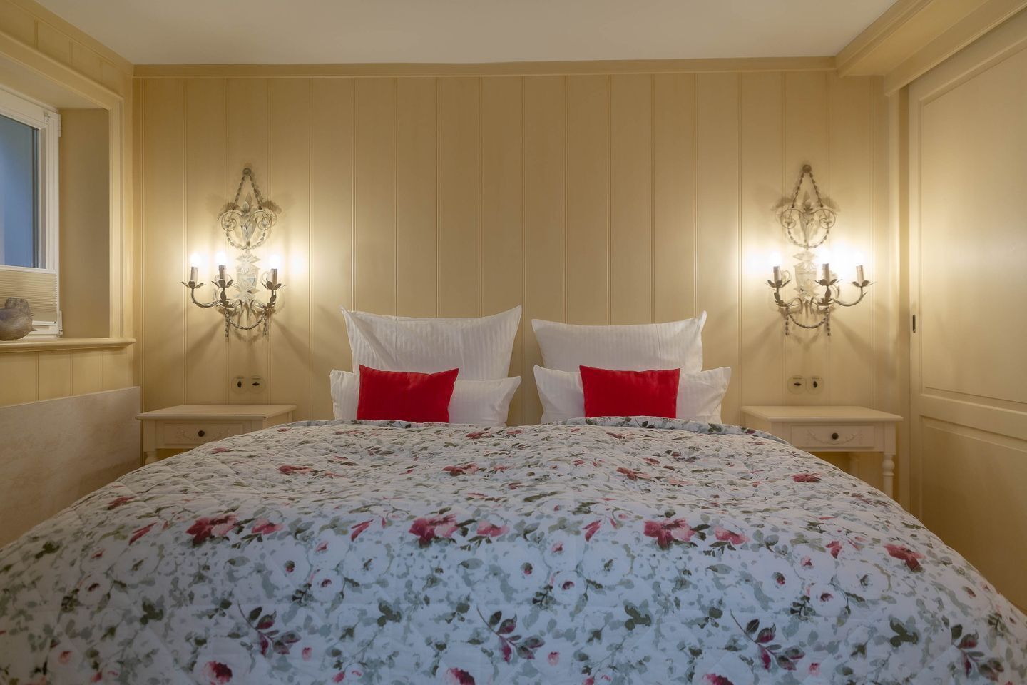 A bedroom with a floral bedspread, two white pillows, red accent pillows, and wall-mounted sconces.