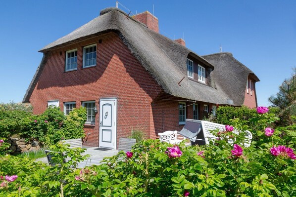 Une maison en briques rouges, avec un toit de chaume, une porte blanche et un jardin où poussent des fleurs roses.