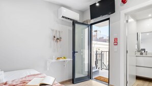 A modern bedroom with a bed, a book, a pink blanket, a glass door leading to a balcony, and a kitchen area. - Seaside Sunset - The Shell by Homie (Calheta)
