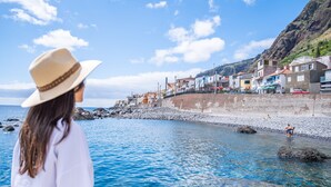 A coastal town with buildings along the shore, a clear sky, and rocky terrain. - Seaside Sunset - The Shell by Homie (Calheta)