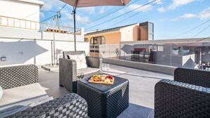 A rooftop terrace with wicker furniture, a table with a fruit platter, and a large umbrella. - Seaside Sunset - The Shell by Homie (Calheta)