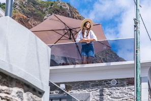 A balcony with a stone wall, a large umbrella, and a mountainous backdrop. - Seaside Sunset - Fisherman s House by Homie (Calheta)