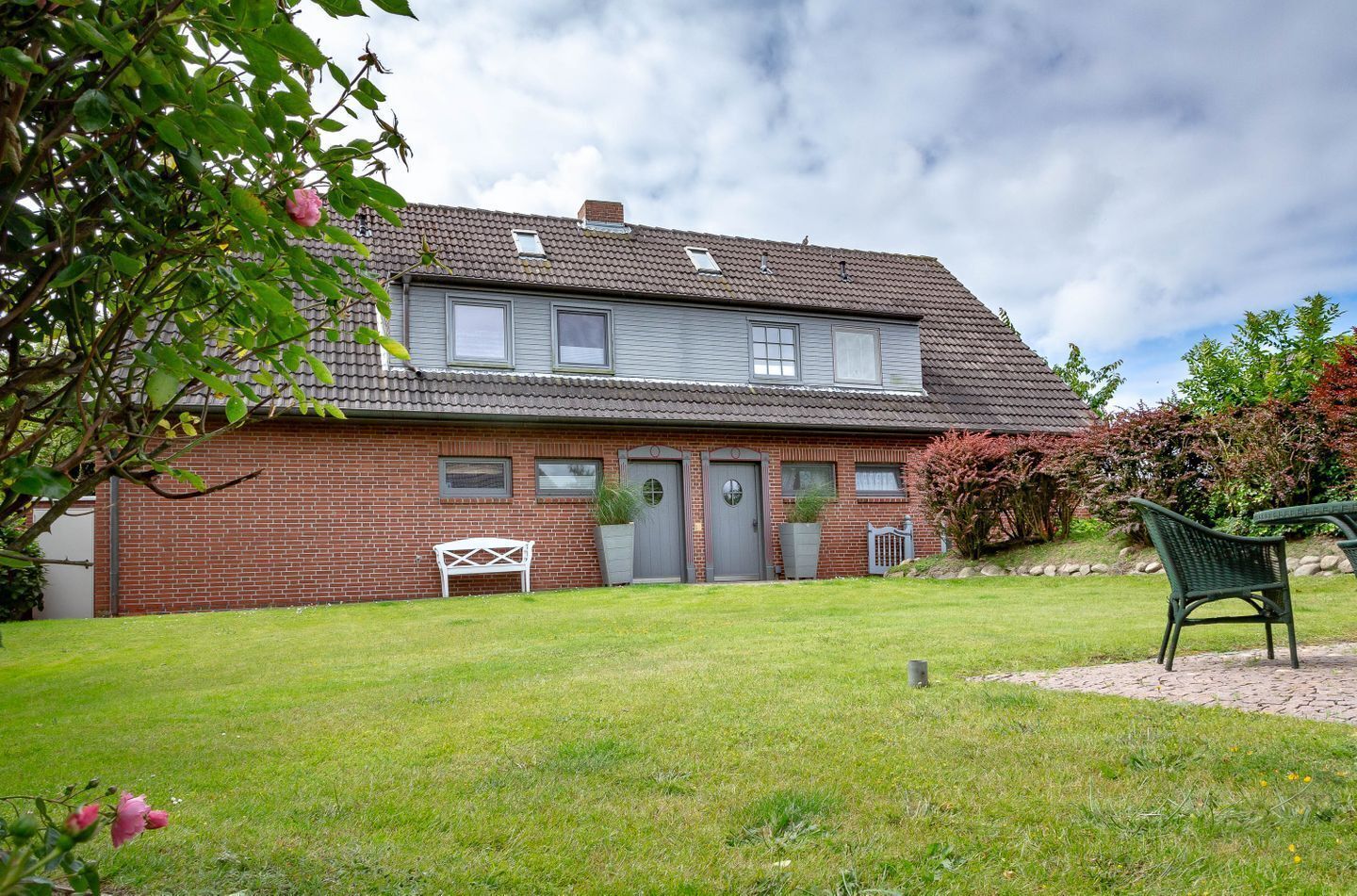 Ein zweistöckiges Backsteinhäuschen mit Ziegeldach, einer Haustür und einem kleinen Garten mit Bank und Stuhl.