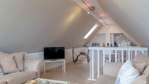 A modern loft apartment with a high ceiling, a flat-screen TV on a white table, a glass coffee table, and a white railing in the background. - Biberhüs OG - Ferienwohnung "biberhüs OG" in Archsum für bis zu 4 Personen (Archsum)