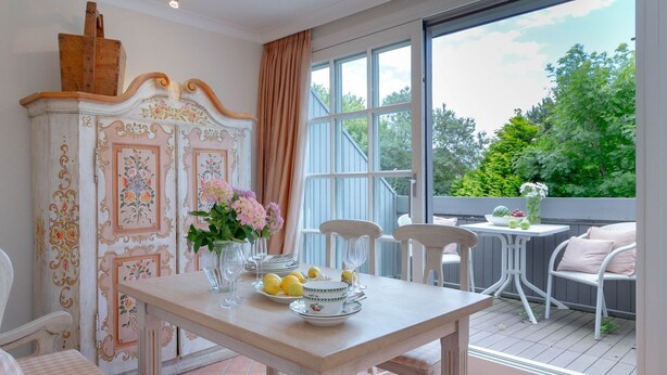 A dining area with a table set for four, a white cabinet with floral designs, and a view of greenery through large windows. - Biberhüs OG - Ferienwohnung "biberhüs OG" in Archsum für bis zu 4 Personen (Archsum)