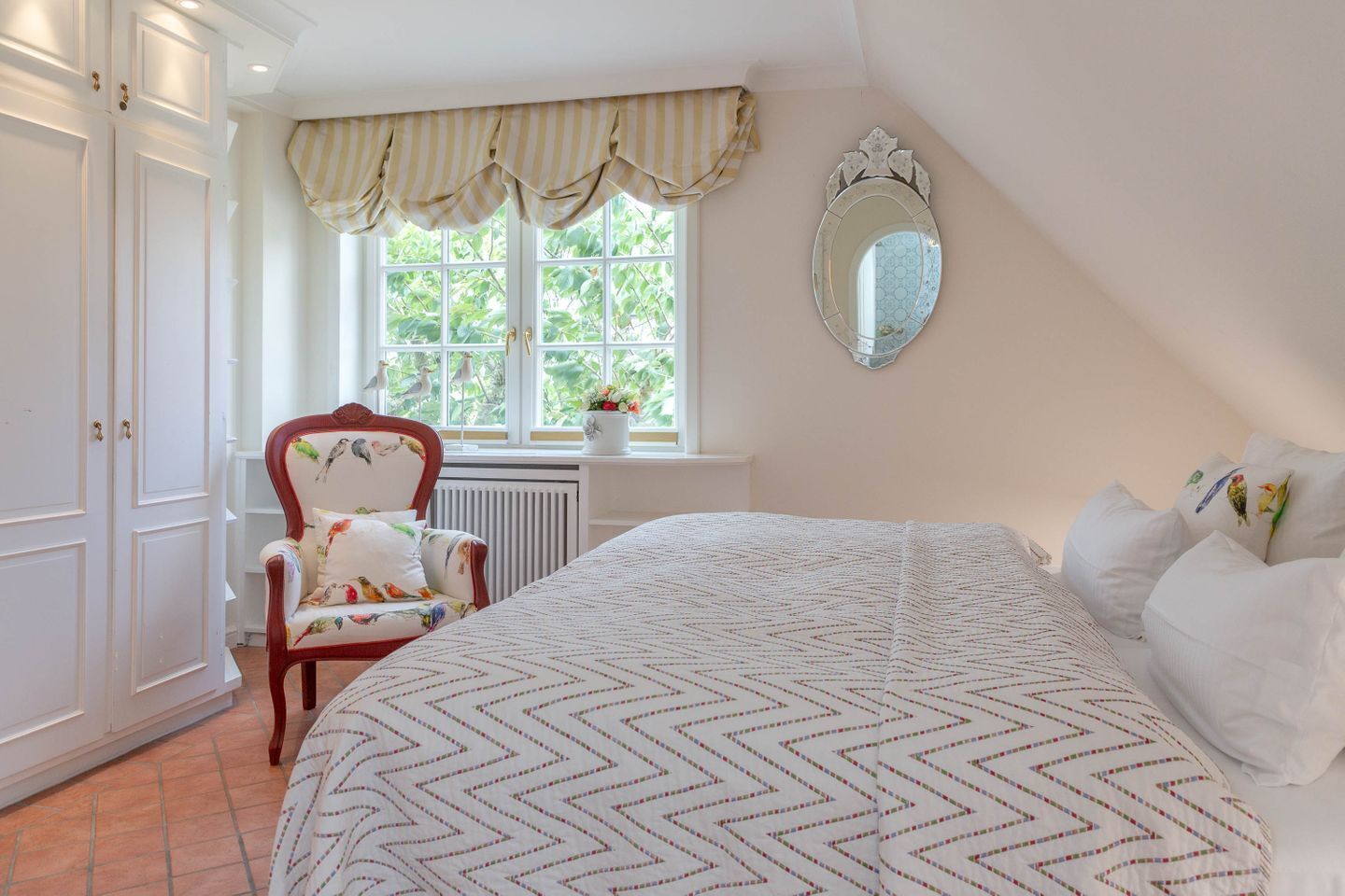 A bedroom with a large bed, a chair with floral upholstery, a window with a view of greenery, and a wall-mounted mirror.